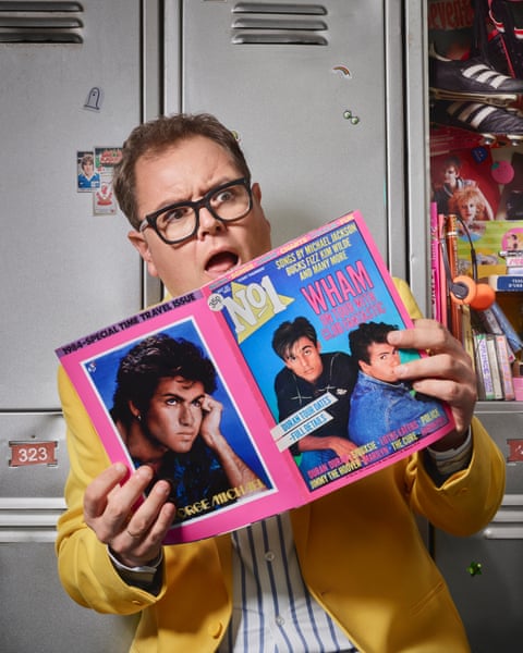Portrait of Alan Carr in front of a school locker holding an 80s style music magazine