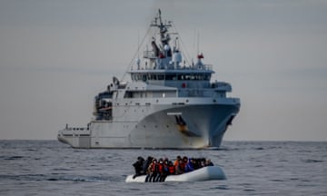 A group of people in an inflatable dinghy at sea with a warship behind them