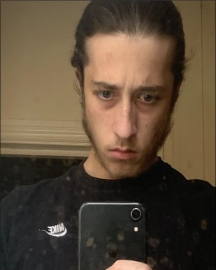 Alfie Coleman looking at a phone; he has light brown hair tied back off his forehead and a thin beard, and wears a black Nike top.