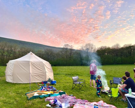 A summer’s evening with blue sky and pink clouds above a green field with a tent, a selection of picnic blankets and a family enjoying the sunset.