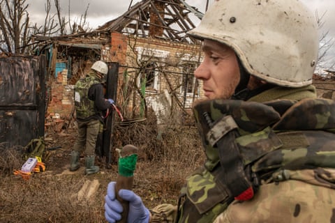 Two people in helmets and camouflage flak jackets search an area next to a damaged home