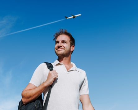 A young man with a backpack on his shoulder smiles as a plane flies overhead.