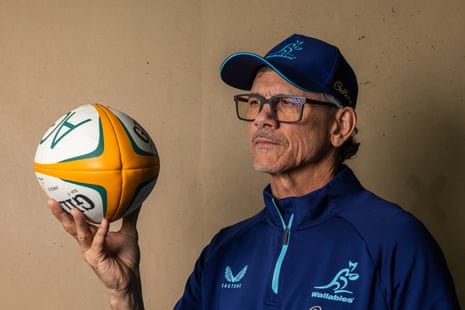 Les Kiss hold a green, gold and white rugby ball in front of his face while wearing a blue Wallabies jumper and cap