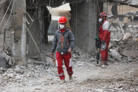 Two workers in all red uniforms and red hardhats walk through the grey rubble and debris of a destroyed building. Both workers have white masks over their faces.