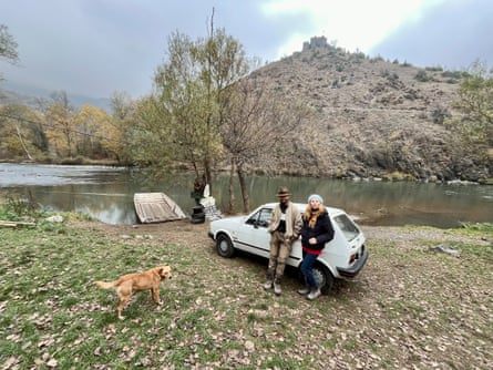 A man and a woman leaning against a small white car by a lake with a raft in it, with a hill behind with a castle on top