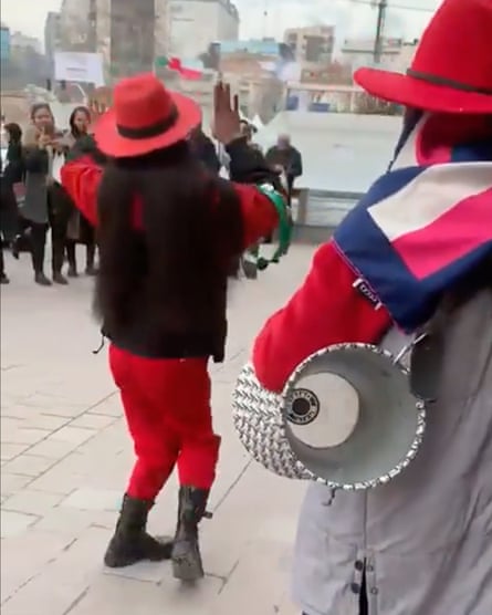 Two women wearing red hats and carrying a drum and a tambourine move in front of people taking pictures.