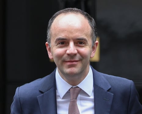Secretary of State James Murray in a blue suit smiles at the camera in front of a dark doorway