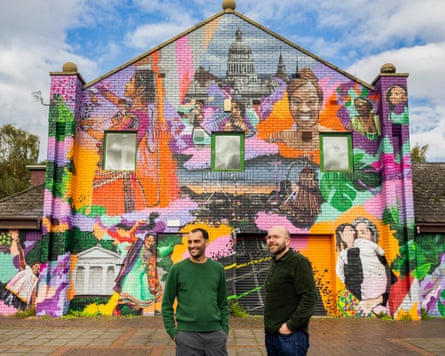 Two people in front of the front wall of a two-storey brick building front painted with a mural