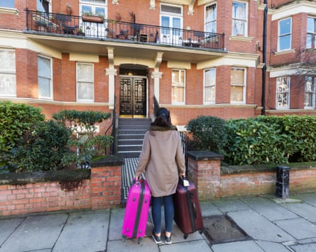 Young person wearing a brown coat and jeans with suitcases outside a house.
