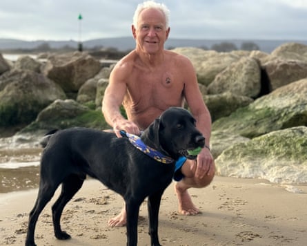 David Howarth shirtless and squatting on his haunches next to his black labrador Beau against a backdrop of beachside boulders
