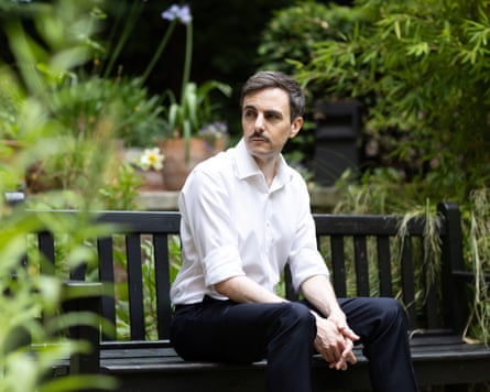 he is in his mid-40s with neat dark hair and moustache and sits on a bench surrounded by greenery. He wears a white open-necked shirt and dark trousers.