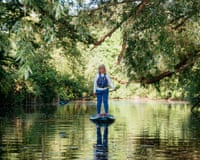 A woman paddleboards on a calm river beneath overhanging trees with dappled sunlight