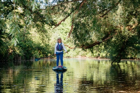 A woman paddleboards on a calm river beneath overhanging trees with dappled sunlight