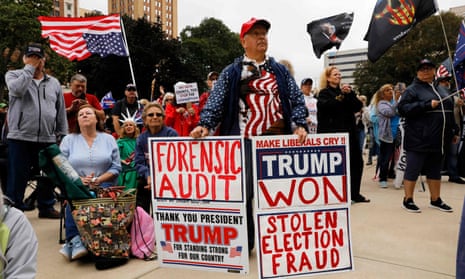 A man wearing a shirt and jacket with American flag designs on it stands at a protest holding two signs. One demands a forensic audit of the 2020 US election. The other reads 'Trump won. Stolen election fraud'.