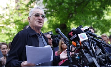 man wearing sunglasses and black jacket holds papers and speaks in front of many microphones