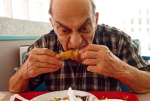 an older man in a checked shirt sits at a table eating fried chicken