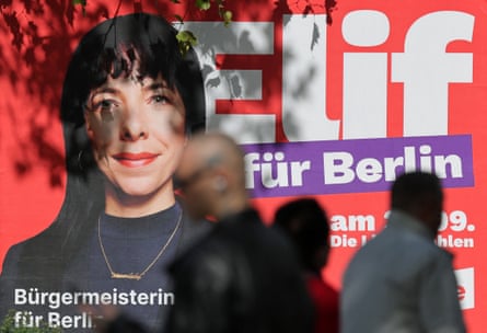 A political campaign poster saying Elif fur Berlin with an image of her face. People walk past the poster in the street.