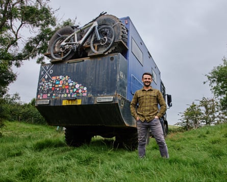 He stands by a huge, battered blue truck with badges stuck on and a bike mounted on the back