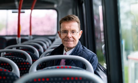 Andy Street, in a quilted jacket over a shirt and tie, sits on a bus in Birmingham, smiling broadly.