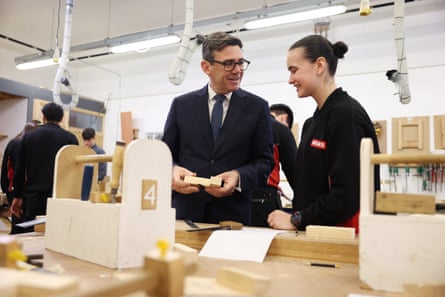 Andy Burnham holds a wooden block as he chats with a smiling apprentice in a carpentry workshop