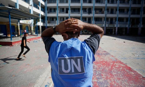 A man wearing a UN blue vest faces away from the camera with his hands on his head in a yard