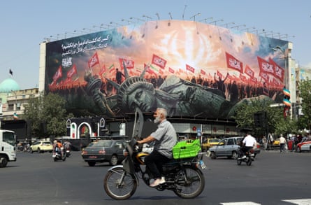 Cars and bikes ride past a big billboard showing the Statue of Liberty knocked down as hundreds of Iranians wave flags behind it