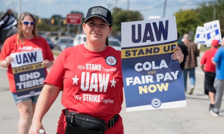 A white woman wearing a red T-shirt with white lettering, among others around her, holds up with one hand a blue, yellow and white sign that says ‘UAW Stand Up COLA and Fair Pay Now.’