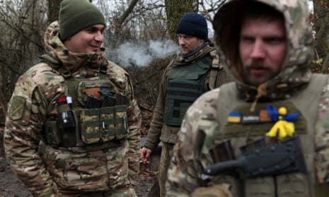 Three Ukrainian servicemen, one of whom is smoking a cigarette, stand in a wooded area