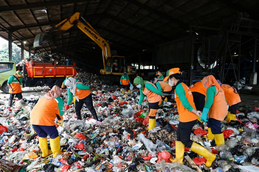 Women in masks, orange uniforms and rubber boots, move through a huge landfill