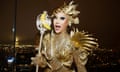 The drag queen Nymphia Wind dressed in a gold outfit holding a stick with a gold sequinned stick with banana at the top