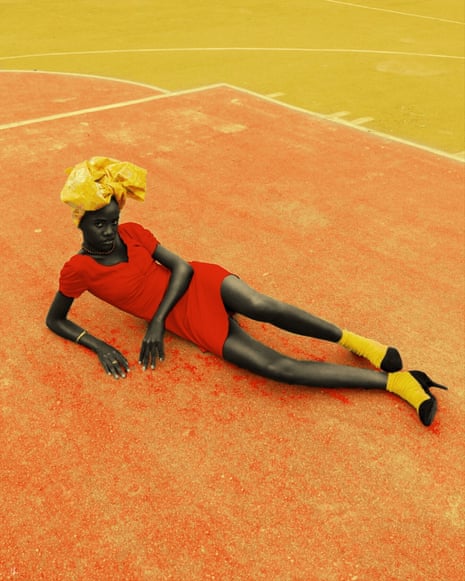 A woman in a bright red dress, yellow headscarf and socks, and black stilettos, lying on a bright orange stadium track