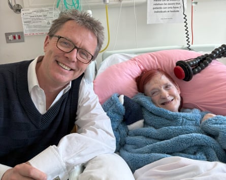 Nicky Campbell smiles with Sian, who is lying under a blue fluffy blanket in what appears to be a hospital bed or a bed in a care facility; she has bright red hair and lies against a pink pillow; he is wearing a white shirt and black sleeveless jumper.