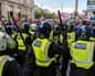 Police officers in riot gear with helmets and batons raised face a large crowd with flags.