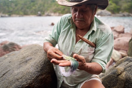 A man holds a skein of yellow-green yarn, sitting on rocks by the sea