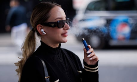 A young woman wearing sunglasses vapes while she walks in the street.