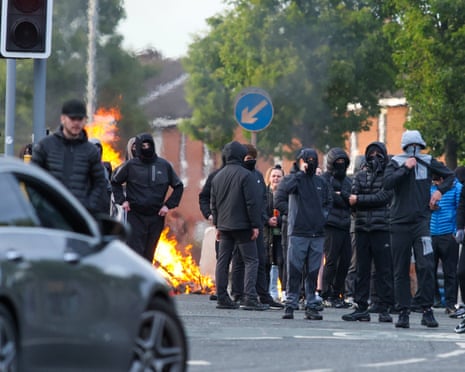 A group of protesters in front of a fire in the street, with dark clothes and covered faces