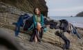 A middel-aged white woman sits on a rock by the sea, wearing a wetsuit and surrounded by other wetsuits