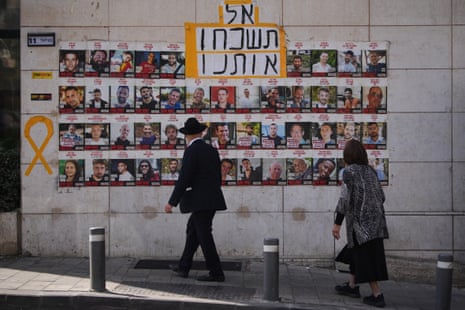 A man in traditional Jewish clothing and a woman walk by, looking at posters on a wall along a street.
