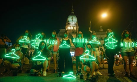 In the dark, a group of young women wearing green face masks stand and kneel in front of a domed government building, holding neon (or lighted) images of coat hangers, candles circled with barbed wire, and the word "Adios."