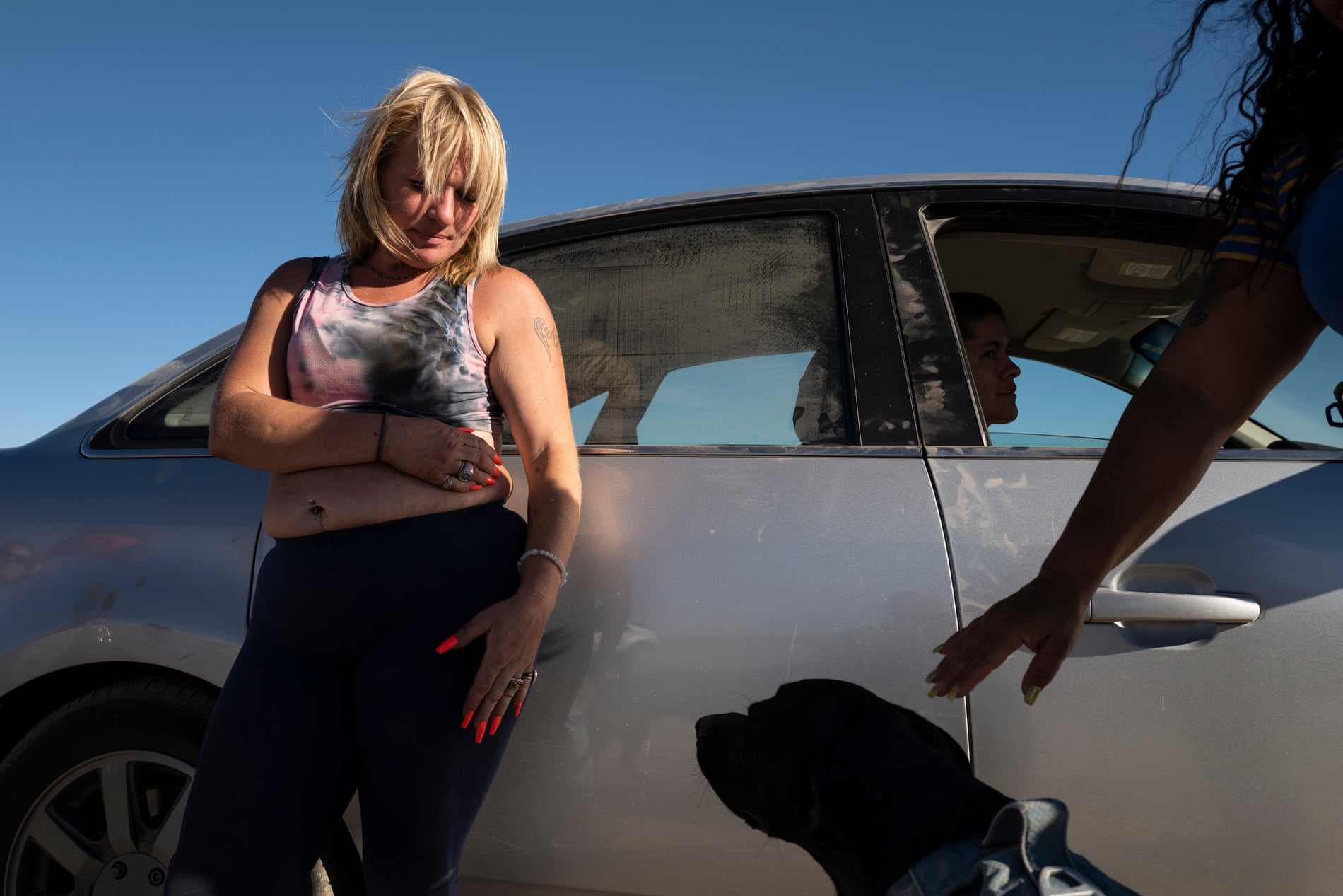 A woman with pink nails stands in front of a dust-covered silver car and looks down at a black dog sitting nearby.