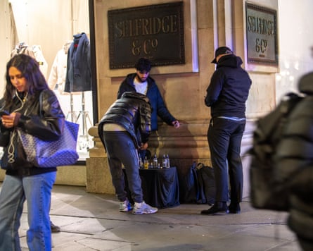 Three men set up table with bottles on it underneath Selfridge & Co signs