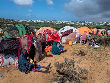Women and children sitting by small crude huts of sticks, cloth and plastic