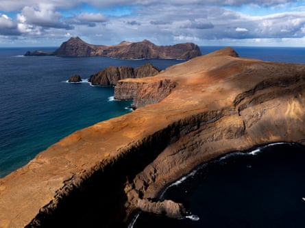 A reddish rocky outcrop extending out to sea towards a small rocky island.