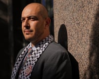 A man with a keffiyeh around his neck stands in dramatic sun and shadow against a wall.