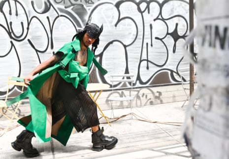 A man dressed in avant-garde clothes walks in front of a graffitied wall.