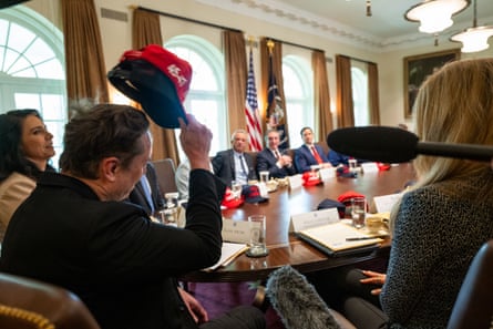 a man seated with others at a table raises his cap