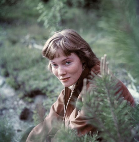 A young woman with short dark blonde hair and wearing a tan jacket crouches amid foliage, looking at the camera and smiling slightly.