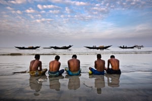 Five fishermen sit cross-legged in shallow water, facing out to sea with bowed heads with four fishing boats in the distance