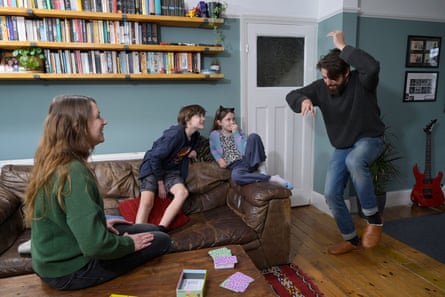 A family of father, mother, son and daughter sit in their front room with a board game on the coffee table. The father is acting out something as the others watch