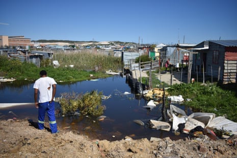 A man stands by a stagnant pool surrounded by corrugated-iron shacks